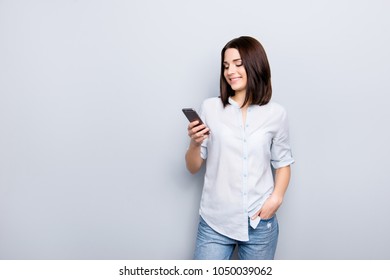 Portrait of pretty, charming, stylish, modern girl in shirt, denim outfit holding hand in pocket of jeans, having smart phone, checking email, searching, using 3G internet, standing on grey background - Shutterstock ID 1050039062