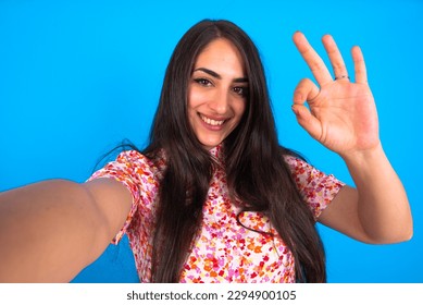 Portrait of pretty cheerful beautiful brunette woman wearing floral dress over blue background make selfie show okey symbol