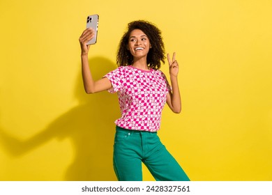 Portrait of pretty cheerful lady hold smart phone make selfie demonstrate v-sign isolated on yellow color background