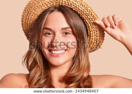 Portrait of pretty cheerful woman wearing straw hat, attractive female looking smiling directly at camera, expresses happyness, keeps hand on hat, being in good mood, posing with bare shoulders.