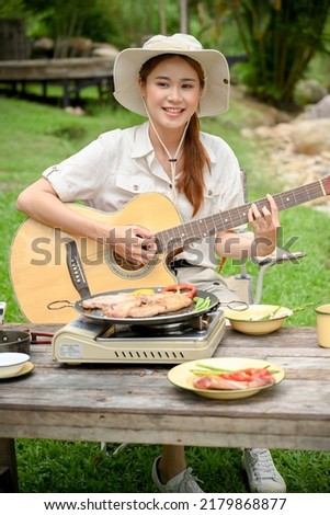 Portrait of a pretty and cheerful young Asian female camper traveler playing her acoustic guitar while camping with her friends at the campground.