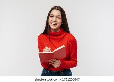 Portrait Of A Pretty Cheerful Young Brunette Woman Wearing Casual Clothing Isolated Over White Background, Taking Notes In A Diary