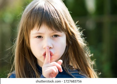 Portrait Of Pretty Child Girl With Gray Eyes And Long Hair Holding Point Finger On Her Lips With Hush Sigh. Cute Female Kid On Warm Summer Day Outside.