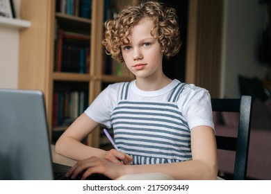 Portrait Of Pretty Curly Schoolgirl Studying With Book And Laptop Preparing For Exam Writing Essay Doing Homework At Home, Looking At Camera. Little Child Student Learning Assignment Making Notes.