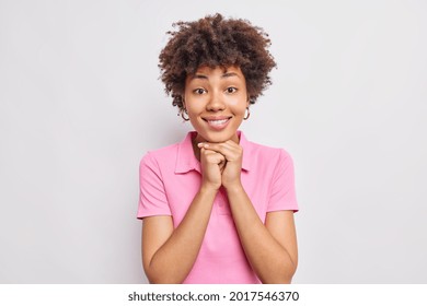 Portrait of pretty dark skinned woman keeps hands under chin smiles pleasantly dressed in casual pink t shirt isolated over white background. Face without makeup. Authentic emotions concept.
