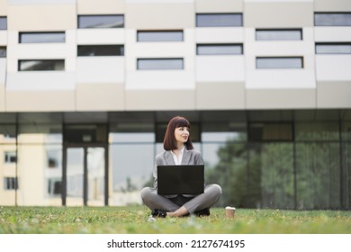 Portrait of pretty excited woman in business suit, sitting crosslegged on green grass in front of office, using a laptop, celebrating success, screaming and raising arms. Well done, good news