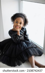 Portrait Of Pretty Female Kid In Black Stylish Dress Sitting Near Window In White Studio And Looking At Camera. Cute Face Of African American Petite Ballerina.