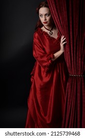  portrait of pretty female model with red hair wearing glamorous historical victorian red ballgown.  Posing with a moody dark background, sitting on  ornate chair.
