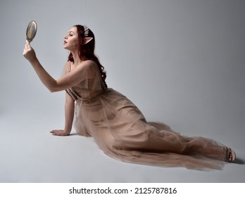 portrait of pretty female model with red hair wearing glamorous fantasy tulle gown and crown.  Posing with gestural arms  holding a hand mirror on a studio background