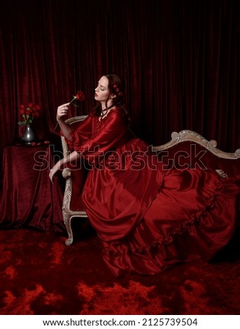  portrait of pretty female model with red hair wearing glamorous historical victorian red ballgown.  Posing with a moody dark background, sitting on  ornate chair.