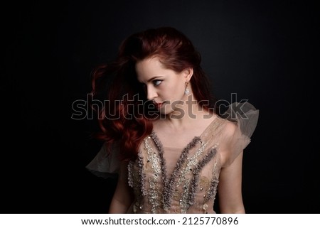  portrait of pretty female model with red hair wearing glamorous fantasy tulle gown and crown.  Posing with a moody dark background.