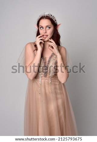 portrait of pretty female model with red hair wearing glamorous fantasy tulle gown and crown.  Posing with gestural arms on a studio background