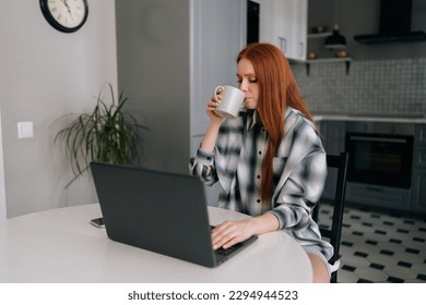Portrait of pretty freelancer female enjoying cup of coffee while remote working or studying on laptop computer sitting at table on kitchen interior. Cute redhead young woman using laptop in morning.