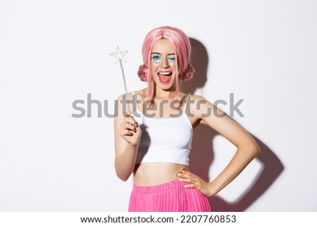 Portrait of pretty girl celebrating halloween in fairy costume, wearing pink wig and bright makeup, holding magic want and smiling, standing over white background