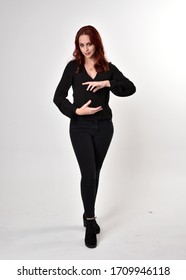 Portrait of a pretty girl with red hair wearing black jeans, boots and a blouse.  full length standing pose, facing the camera with a on a studio background.