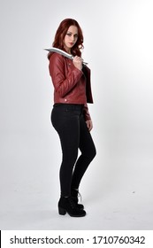 Portrait of a pretty girl with red hair wearing black jeans and boots with leather jacket.  full length standing pose holding knife a studio background.
