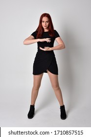 Portrait of a pretty girl with red hair wearing short black dress and boots, full length standing pose on a studio background.