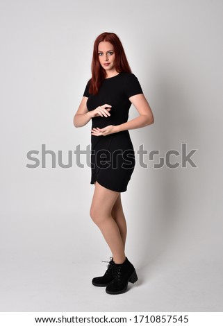 Portrait of a pretty girl with red hair wearing short black dress and boots, full length standing pose on a studio background.