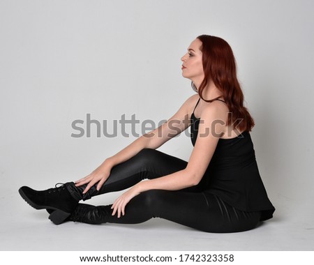  portrait of a pretty girl with red hair wearing black leather pants and top. Full length sitting pose isolated against a  grey studio background
