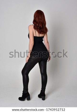 Portrait of a pretty girl with red hair wearing black leather pants, top and boots.  full length standing pose with back to the camera, isolated against a studio background