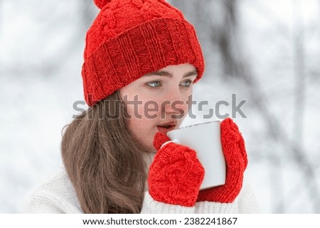 Portrait of pretty girl in red hat and mittens in snowy park. Cute young woman drink hot beverage from cup in winter outdoor
