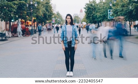 Portrait of pretty girl in trendy clothing looking at camera standing on pedestrian street by herself when busy men and women are moving around in haste.
