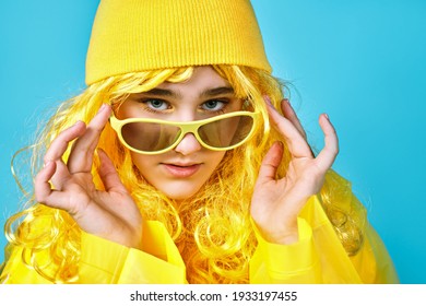 portrait of pretty girl in a yellow hair hat. teenager in cap and raincoat and with glasses in his hands. blue background