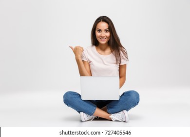 Portrait of a pretty happy asian girl pointing finger away at copy space while sitting and working on laptop computer isolated over white background