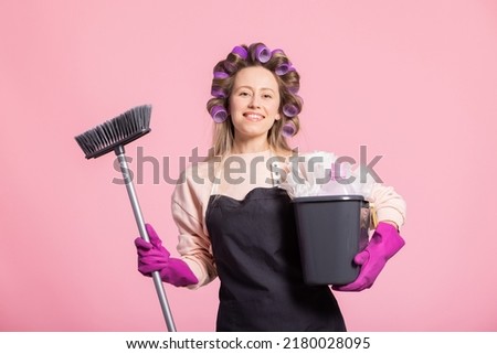 Portrait of pretty housewife good mood wife holds cleaning equipment bright pink background woman wears latex gloves pink apron and rollers on head.