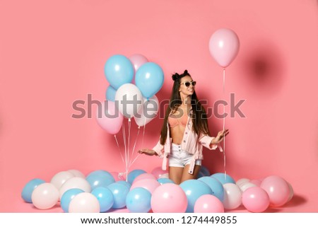 Portrait of pretty joyful young woman in blue dress holding and hugging red box with gift present on pastel pink background with colorful air balloons. Birthday holiday party, people sincere emotions