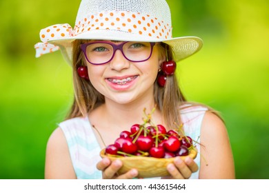Portrait of Pretty little child with full bowl of fresh cherries. Young cute caucasian blond girl with teeth braces and glasses.