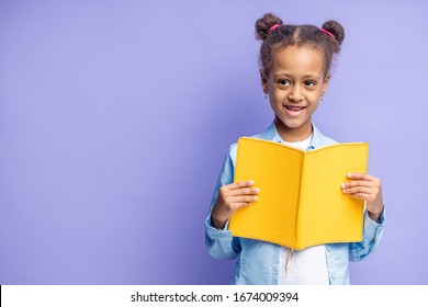 Portrait Of Pretty Little Child Reading Book Isolated Over Purple Background. Happy Girl With Curly Hair Go In 1st Grade At School. Natural Beauty, School, People, Children Concept