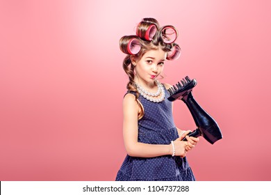 Portrait Of A Pretty Little Girl With Curlers In Her Hair Holding Hair Dryer. Studio Shot Over Pink Background. Kid's Fashion. 