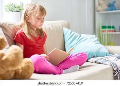 Portrait Of Pretty Little Girl Reading Book Sitting Cross Legged On Comfortable Couch In Cozy Living Room At Home