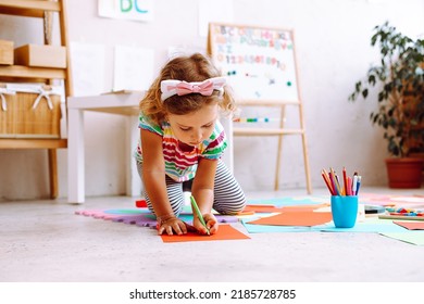 Portrait Of Pretty Little Girl With Short Fair Hair Wearing T-shirt, Trousers Sitting Near Magnetic Board With Numbers, Alphabet, Drawing With Pencil On Red Sheet Of Paper Around Colorful Papers