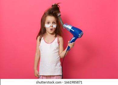 Portrait Of Pretty Little Girl With Tangled Hair Comb In Her Hair, Kid Holding Hair Dryer And Having Astonished Facial Expression, Has Problem, Model Posing Isolated On Pink Background. Kid's Fashion.