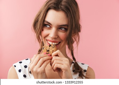 Portrait of a pretty lovely girl wearing dress standing isolated over pink background, eating chocolate chip cookie