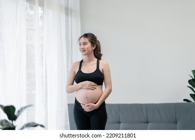 Portrait Pretty Pregnant Asian Woman In Sportswear Is Standing And Rubbing Her Tummy With While Looking Through The Window. Concept Healthy Pregnancy.