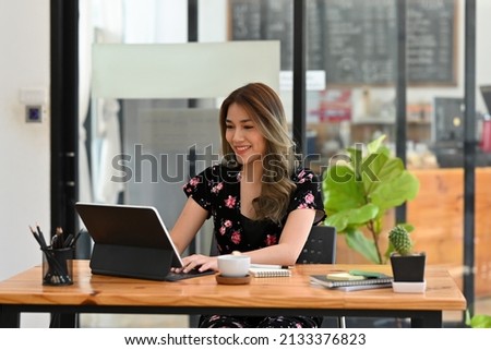 A portrait of a pretty smiley Asian woman sitting in the office looking at a tablet and working on a document, for business and technology concept. 
