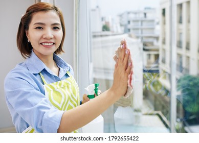 Portrait of pretty smiling Asian housewife wiping apartment window and looking at camera