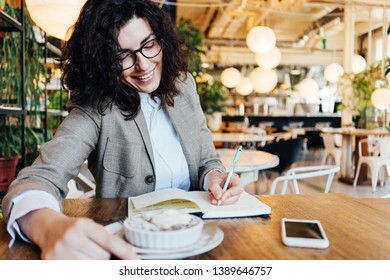 Portrait Of Pretty Smiling Business Lady Sitting In Eco Cafe With Notepad, Phone, Cup Of Coffee, Writing Down Ideas With A Pen