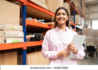Portrait of pretty smiling female employee holding inventory clipboard while standing against rack at warehouse