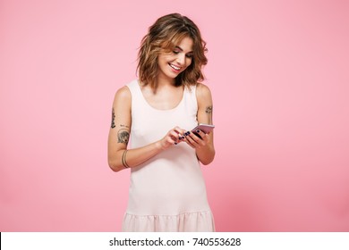 Portrait of a pretty smiling girl in summer clothes using mobile phone while standing isolated over pink background - Powered by Shutterstock