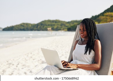 Portrait Of Pretty Smiling Young Black Woman Resting On Chaise-lounge On Sandy Beach And Working On Laptop