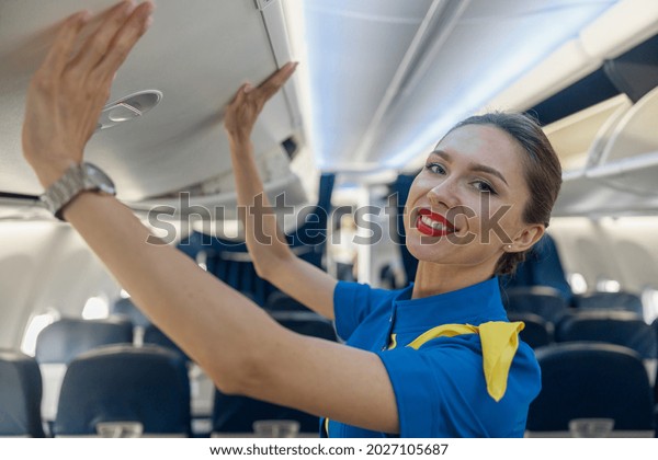 Portrait of\
pretty stewardess in blue uniform smiling at camera while closing\
hand luggage compartment, staying on the aisle inside the plane.\
Transportation, occupation\
concept