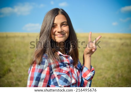 portrait of pretty teenage girl having fun showing peace sign happy smiling & looking at camera on green field under blue sky summer outdoors copy space background