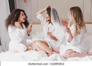 Portrait of pretty three women 20s celebrating bachelorette party and drinking glasses of champagne in luxuty apartment or hotel room while bride trying on wedding veil