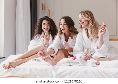 Portrait Of Pretty Three Women 20s Wearing Bathrobe Sitting In Big Bed In Posh Apartment Or Hotel Room And Painting Nails During Hen Party