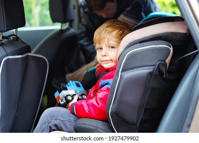 Portrait Of Pretty Toddler Boy Sitting In Car Seat. Child Transportation Safety. Father Clips On Another Son On Background. Cute Healthy Kid Boy With Toy Happy About Family Vacations With Car