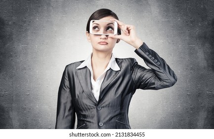 Portrait of pretty woman covering her eyes with smartphone. Businesswoman showing mobile phone with eyes on screen. Corporate businessperson on grey wall background. Mobile communication layout.